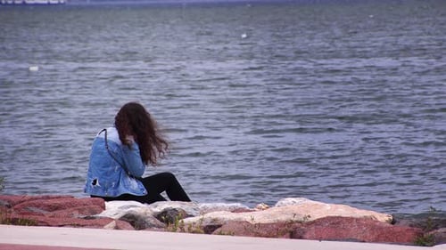Curly Haired Young Girl Sits On Stones By The Sea