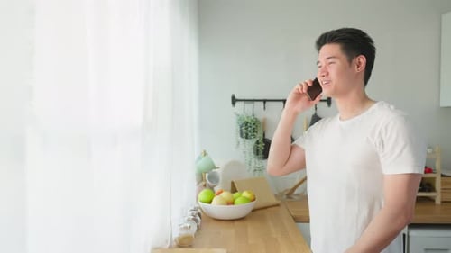 Asian handsome man talking on mobile phone call stand in kitchen at home.