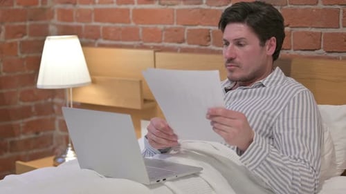 Man Reviews Documents in Bed with Laptop