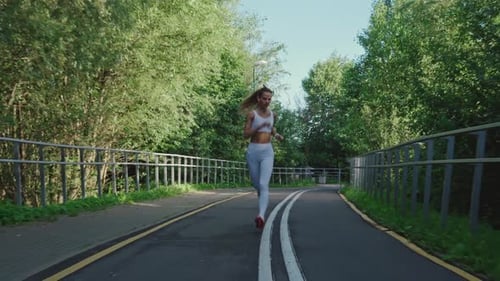 Beautiful Blonde Girl in a White Top and Leggings Jogging in the Park on a Sunny Day