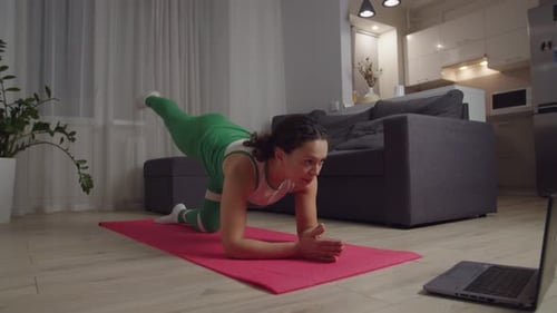 Mujer en ropa deportiva realizando ejercicios de estiramiento de piernas con computadora portátil en interiores