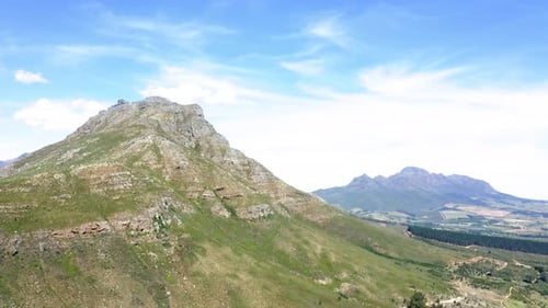 Aerial drone passing close-up of green mountain peak with forest and mountain range in background, s