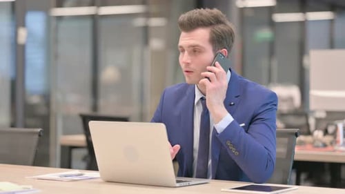 Businessman Talking on Phone While Using Laptop in Office