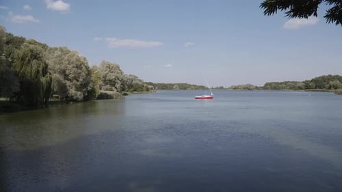 Segelboot mit rotem Rumpf steht mit abgesenkten Segeln in der Mitte des schiffbaren Sees