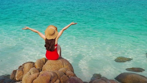young woman sitting with arms raised on stone beach with sea at Koh MunNork Island, Rayong, Thailand