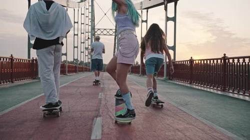 Rear View of Four Young Men Boys and Girls Riding Skateboards Across the Large City Bridge at Dawn