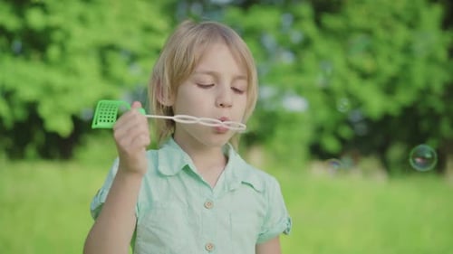 Close-up of Little Caucasian Boy Blowing Soap Bubbles on Sunny Meadow. Cute Blond Child with Green