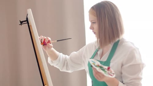 Woman is Intently Drawing with Brush on Canvas Holding Palette in Hand Side View