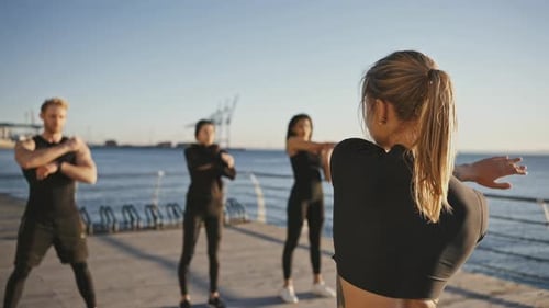 Young Female Coach Showing Exercises to Group of Sporty People Practicing Warm Up Training on Pier