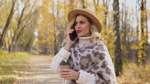 Beautiful Blonde Woman in a Brown Hat Speaks on the Phone in the Autumn Park