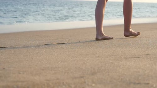 Female Feet Walking on Golden Sand at the Beach with Ocean Waves at Background. Legs of Young Woman