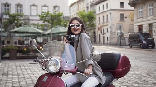 Junge Frau in stilvoller Brille hält Vintage-Kamera in der Hand und sitzt auf dem modernen Roller