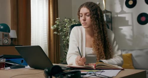 Girl with Curly Hair Sits at Desk in Living Room and Works on Laptop Taking Notes Focused Young