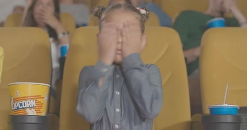 Portrait of Pretty Frightened Little Girl Closing Eyes with Hands in Movie Theater. Charming Cute