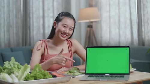 Asian Woman Pointing Green Screen Laptop On The Table And Smiling To Camera
