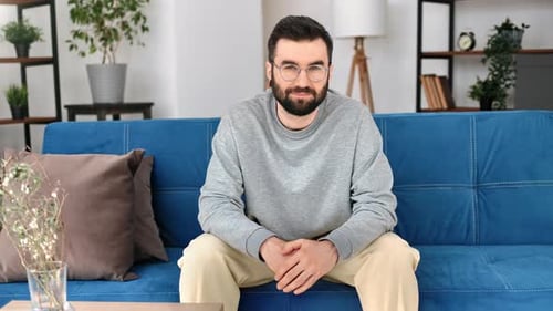 Confident Handsome Hipster Bearded Man in Eyeglasses Sitting on Blue Couch at Cosiness Living Room