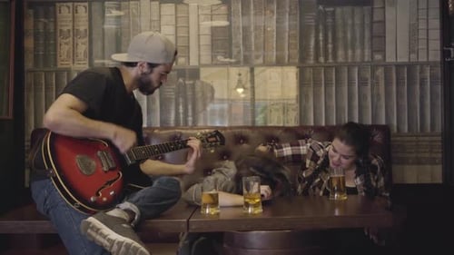 Young Positive Bearded Man Playing Guitar in the Bar, His Friends Sitting Near