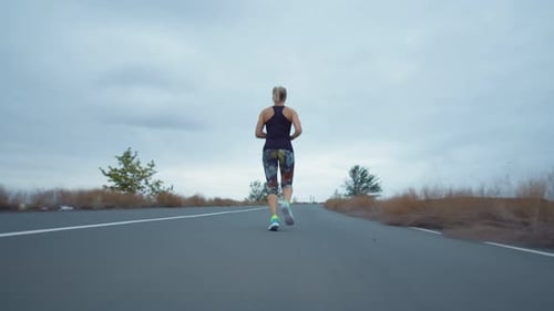 Woman in Sportswear is Jogging Outdoors Background Cloudy Sky Camera Gradually Approaching Back View