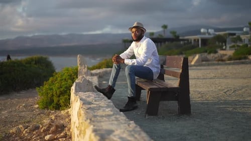 Side View of Thoughtful African American Gay Man Sitting in Sunset Rays on Bench Looking Away
