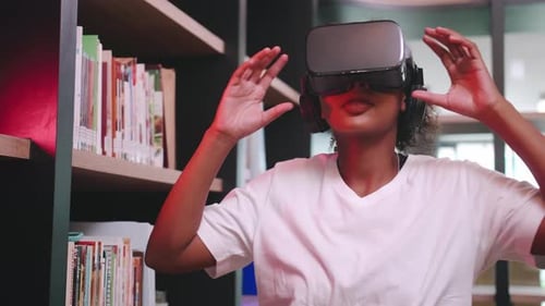 A schoolgirl sitting in front of library's bookshelf and using virtual reality glasses.