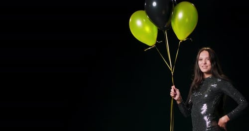 A Woman in a Black Brocade Dress Dances While Holding Black and Gold Balloons Against a Black Studio