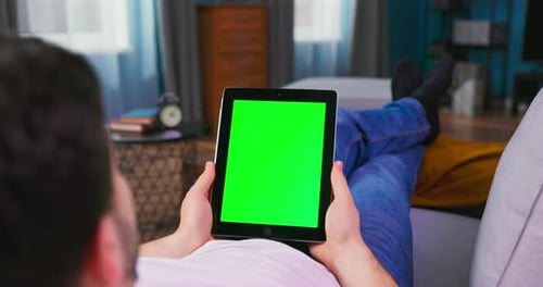 Young Man at Home Laying on a Couch Using with Green Mockup Screen Tablet Computer