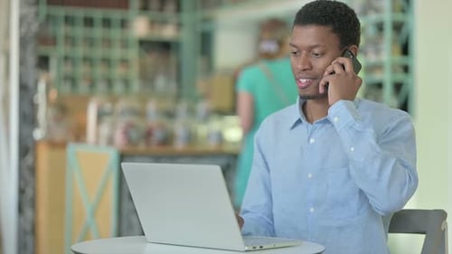 Young African Man with Laptop Talking on Smartphone in Cafe