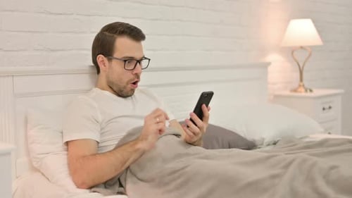 Success Young Man Celebrating on Smartphone in Bed