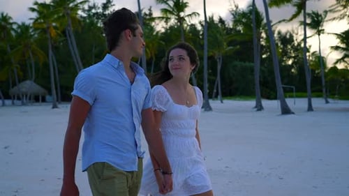 Young couple smiling, walking hand in hand on white sandy beach with palm trees at sunset. Romantic