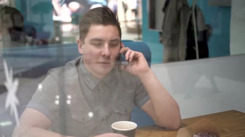 A Young Man Behind the Glass Sits in a Cafe and Speaks on the Phone