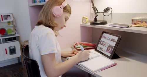 Girl Writes in Notebook at Desk With Tablet