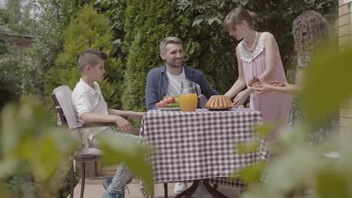Dad, Mom and Their Two Kids Sitting at a Table, Eating Lunch in the Garden Enjoying a Sunny Day