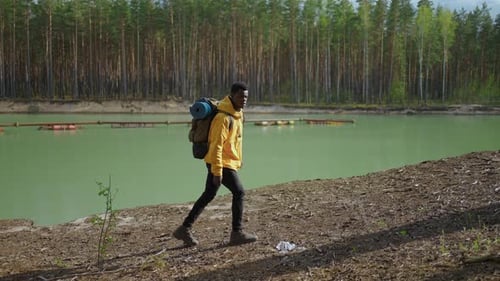 A Young Black Man in a Yellow Cursor Climbs Up with a Backpack Uphill in the Woods Looking at the