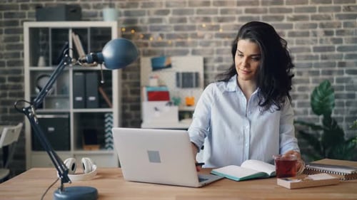 Young Woman Employee Using Laptop and Writing in Notebook at Desk in Office