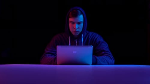 Young Man in a Hood with a Laptop, Hacker Makes a Hack Through a Laptop. Blue and Red Light Falls on