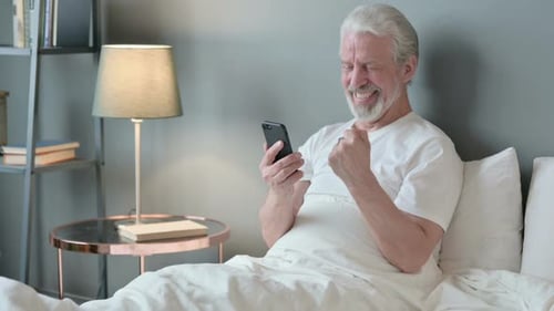 Senior Man Using Phone in Bed Celebrating