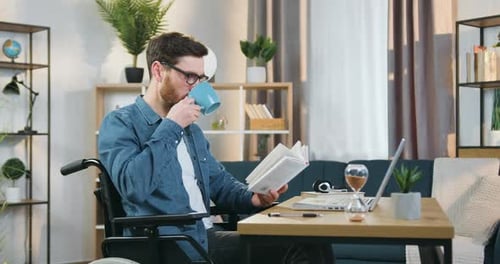 Hombre con gafas sentado en silla de ruedas y leyendo un libro simultáneamente bebiendo té en casa