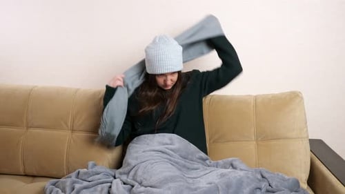 Woman with Plaid Puts on Scarf Sitting on Sofa in Cold Room