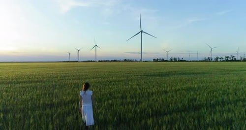 Beautiful girl walking on a green wheat field with windmills for electric power production
