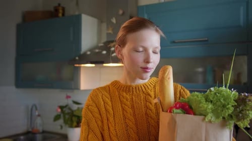 Beautiful Young Woman with Groceries Bag in Hands at Kitchen Looks Camera Smiling