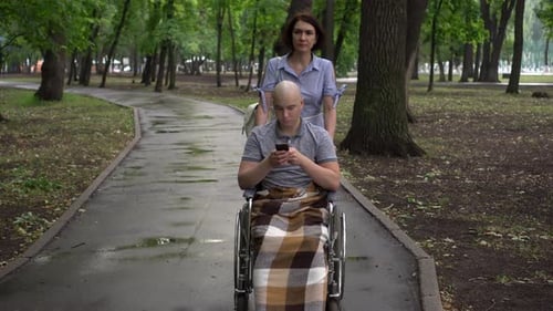 A Woman Is Leading a Young Man with Oncology in a Park in a Wheelchair