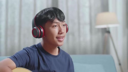 Close Up Of Asian Boy Composer With Notebook On Table Wearing Headphones And Playing Guitar At Home