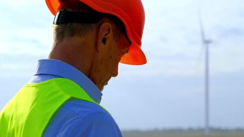 Engineer on Foreground Looks at Blurred Rotating Windmill