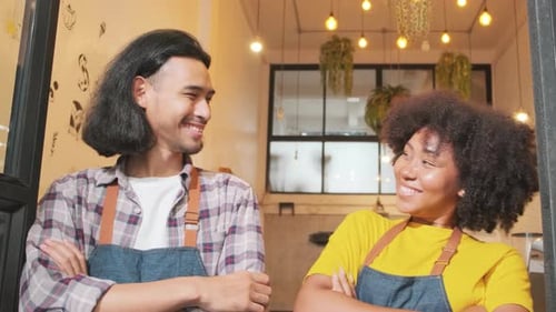 Two baristas at a cafe door, arms crossed, teasing together with a happy smile.