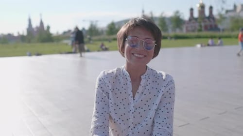 Une femme aux lunettes de soleil colorées est assise sur une scène dans un parc urbain et profite du soleil