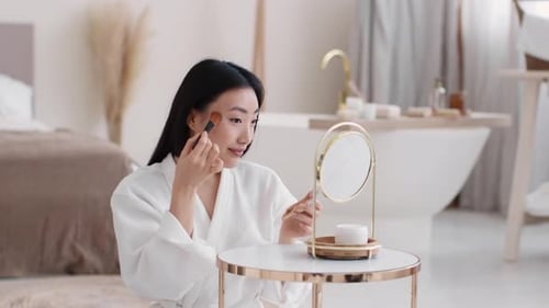 Young Pretty Asian Woman Making Makeup Herself Applying Rouge on Face Looking at Mirror at Bathroom