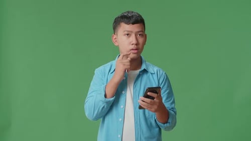 Young Asian Boy Use Mobile Phone And Thinking About Something While Standing On Green Screen