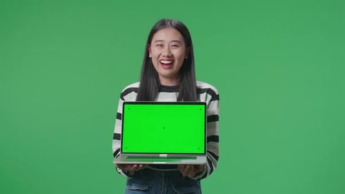 A Smiling Asian Woman Saying Wow While Holding Green Screen Computer In The Green Screen Studio