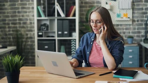 Pretty Young Lady Speaking on Mobile Phone Typing on Laptop in Office