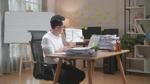 Asian Man With Documents Explaining Work On Paper While Having Video Call On Laptop At The Office